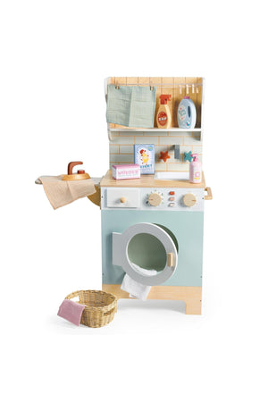 A charming wooden laundrette toy features a pastel blue washing machine, shelves with accessories, and a wicker laundry basket, promoting imaginative play for young children. Ideal for eco-conscious families.