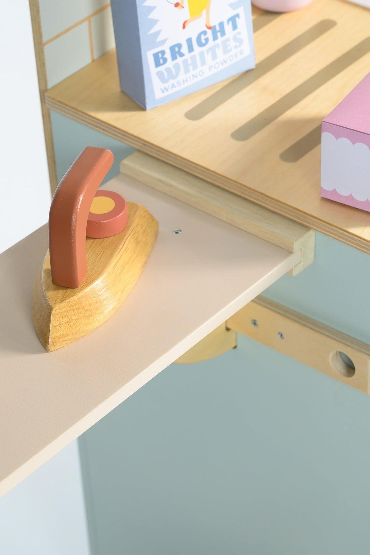 A wooden ironing board features a natural wooden iron and colorful laundry accessories. The light wood and pastel colors create a playful, inviting atmosphere for imaginative play.