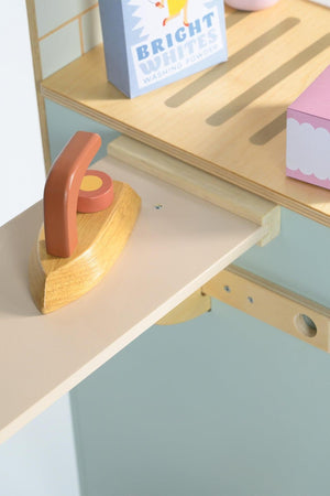A wooden ironing board features a natural wooden iron and colorful laundry accessories. The light wood and pastel colors create a playful, inviting atmosphere for imaginative play.