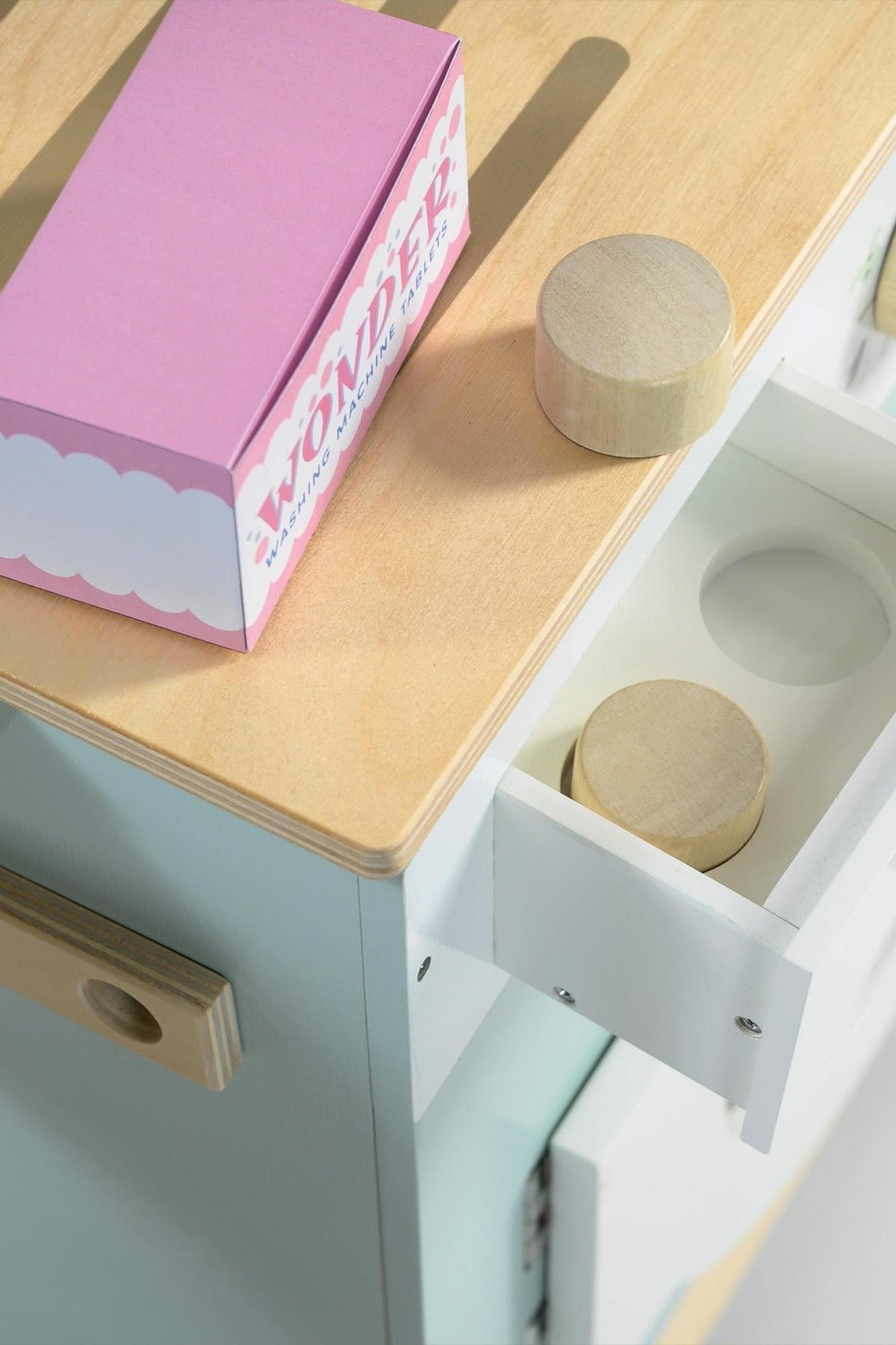 A pastel-colored wooden laundry unit features a smooth light wood countertop and a white drawer, showcasing two round wooden pieces. The design is playful and suitable for childrens imaginative play.