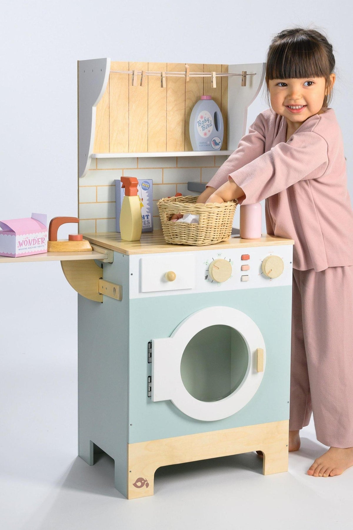 A young child in a pink outfit plays with a wooden toy laundrette, featuring a light blue washing machine, wicker basket, and various pretend cleaning products on a countertop.