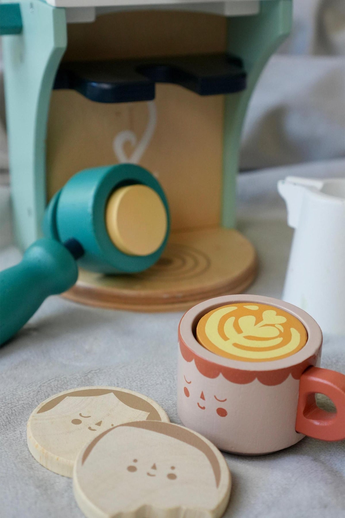 A pastel-colored wooden babyccino maker with a cute bird dial, accompanied by a smiling coffee cup and decorative coasters.