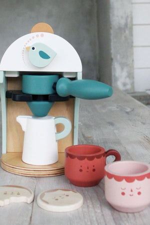 A wooden babyccino maker in soft colors features a bird dial, accompanied by two cute coffee cups and biscuit pieces.