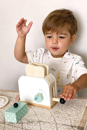 A young child operates a wooden toy toaster with a bird design, surrounded by play food on a table.