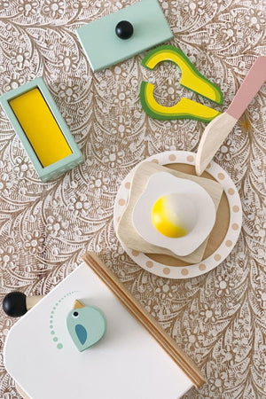 A colorful wooden breakfast playset featuring a poached egg, avocado, a knife, and a butter dish on a floral background.