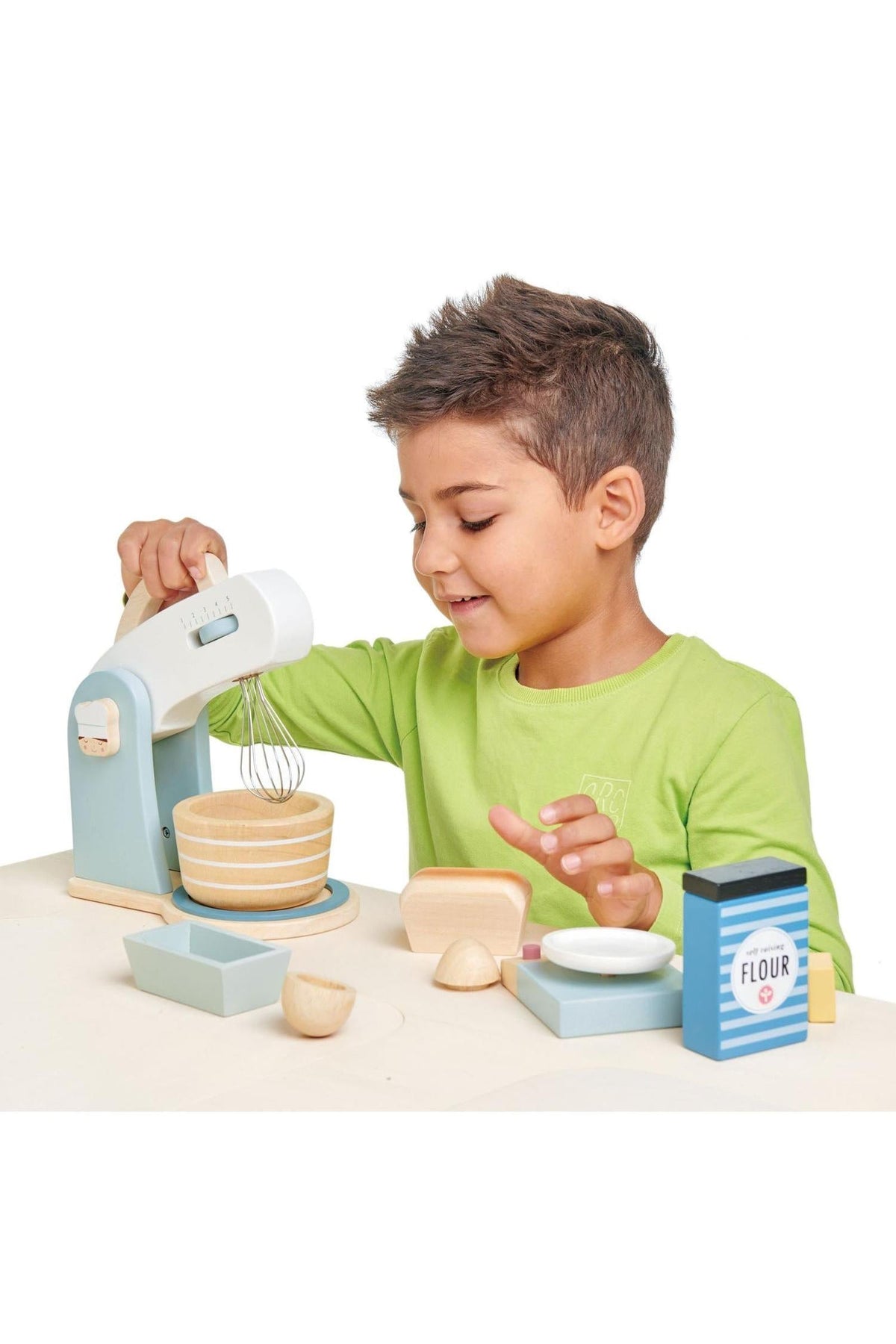 A young boy in a green shirt uses a wooden toy mixer with a bowl, surrounded by colorful baking accessories.