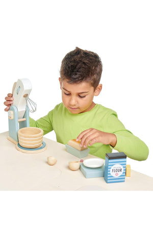 A child in a green shirt uses a wooden mixer and bowls, engaging in imaginative baking play with colorful ingredients.