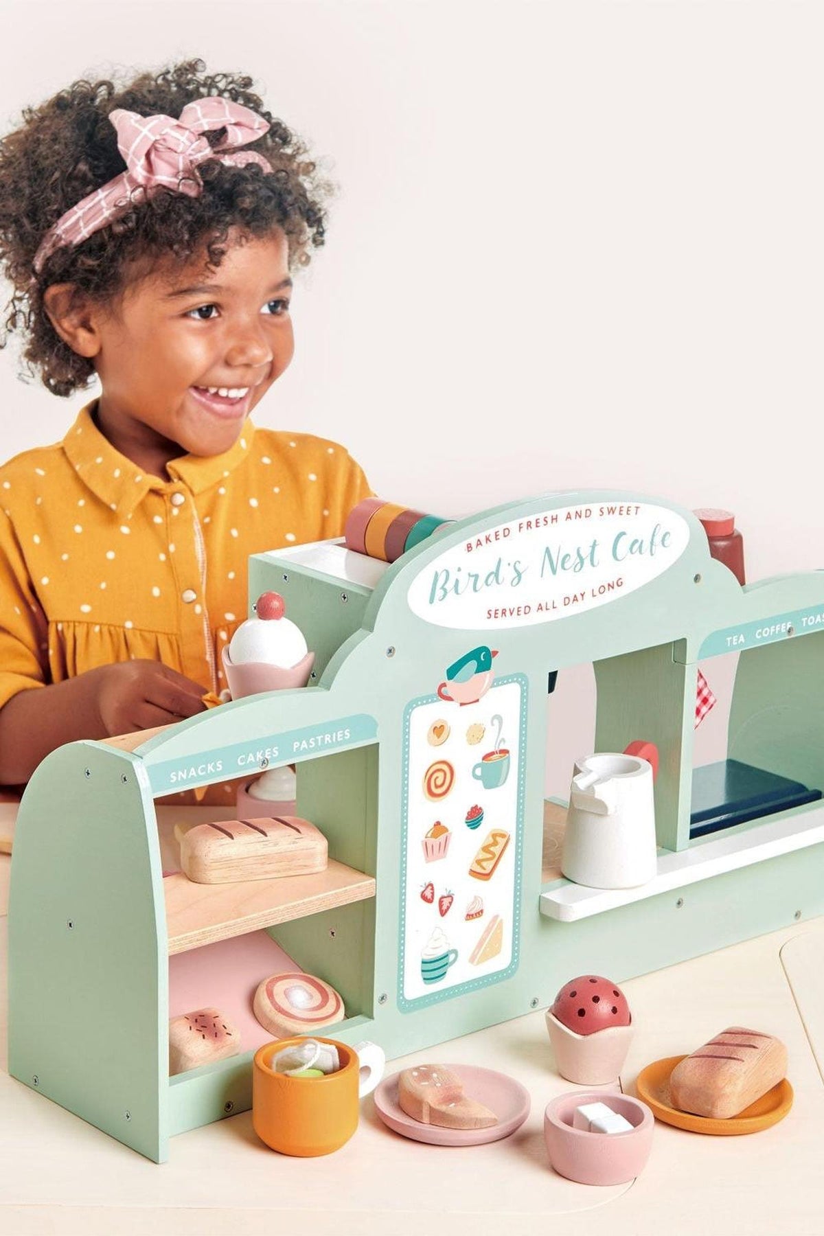 A smiling child plays with the Tender Leaf Birds Nest Café, featuring pastel colors and various pretend food items.