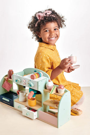 A smiling child in a yellow polka-dot dress holds a toy cup next to a colorful wooden café playset.