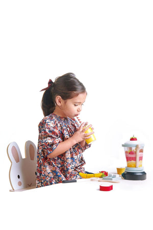 A child drinks from a yellow cup with a straw while playing with a colorful wooden smoothie blender set.