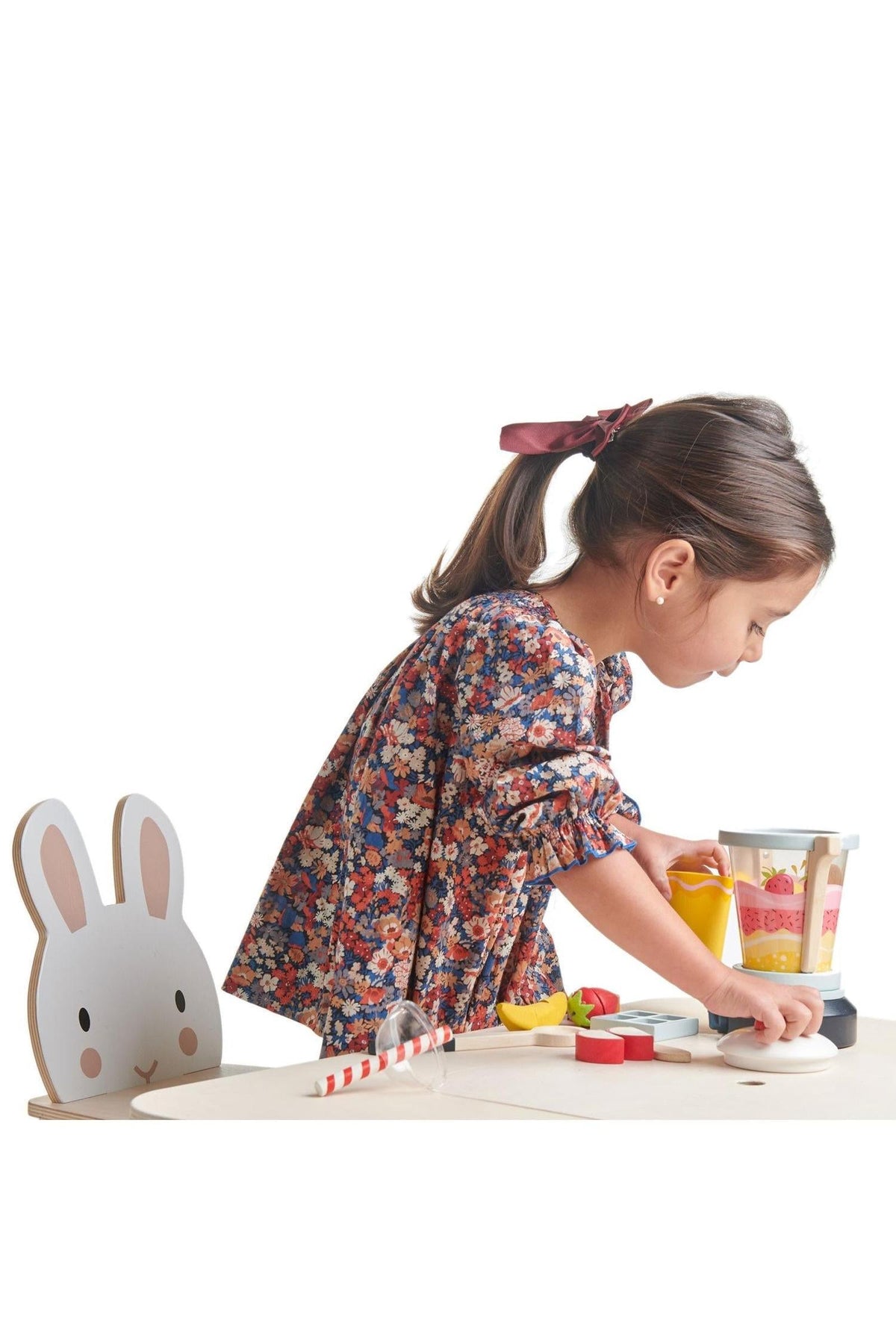 A young girl in a floral dress plays with a colorful wooden toy blender and fruit, set against a white.