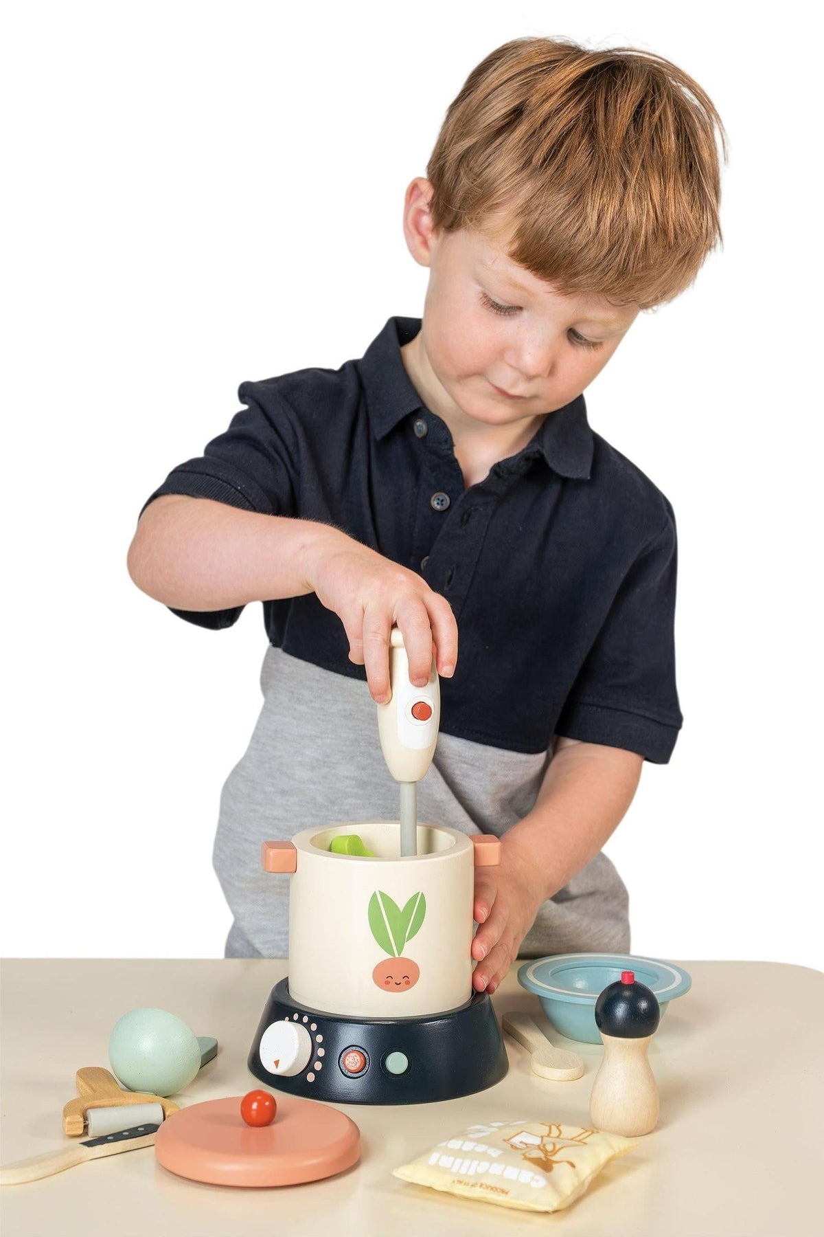 A young child with short, light brown hair is using a white hand mixer in a colorful wooden soup maker.