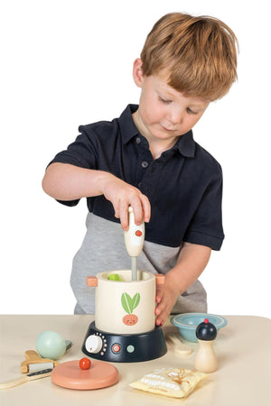 A young child with short, light brown hair is using a white hand mixer in a colorful wooden soup maker.