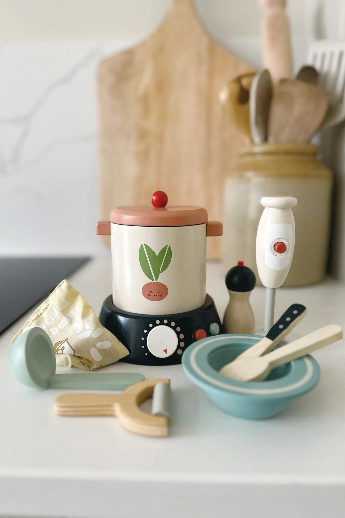 A wooden soup maker set featuring a cream-colored pot with a plant design, alongside utensils like a whisk, bowls, and a striped apron, all arranged on a kitchen countertop.