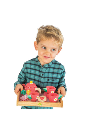 A young child with curly hair holds a wooden tea tray set featuring colorful cups, a teapot, and a biscuit.