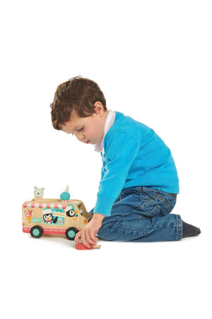 A child in a blue sweater plays with a wooden gelato van toy, featuring penguins and colorful ice cream cones.