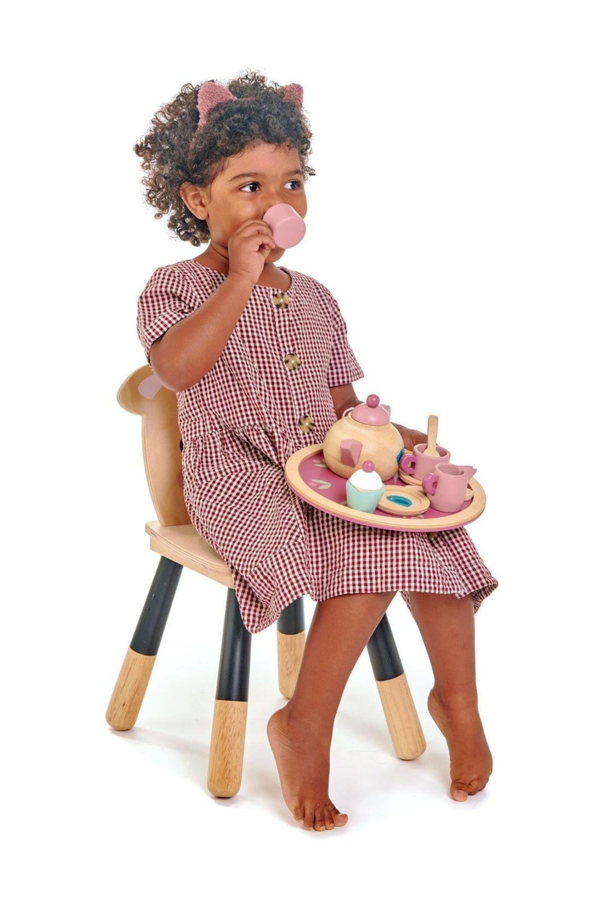 A young girl sits on a wooden chair, sipping from a pink cup while holding a round tray with a wooden tea set, featuring pastel-colored cups and a teapot.