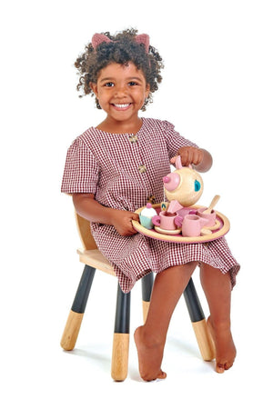 A smiling child sits on a wooden chair, holding a pink wooden teapot and a round tray with colorful tea set pieces.