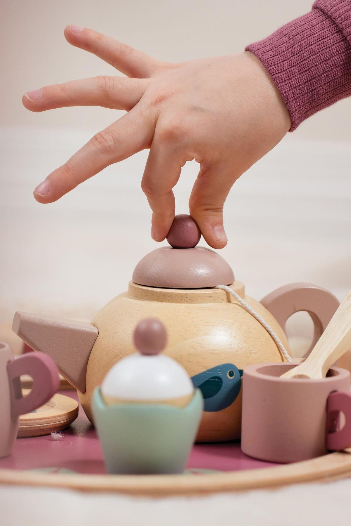 A childs hand delicately places a wooden lid on a light-colored wooden teapot, surrounded by pastel-colored cups, a cupcake, and a milk jug on a round tray.