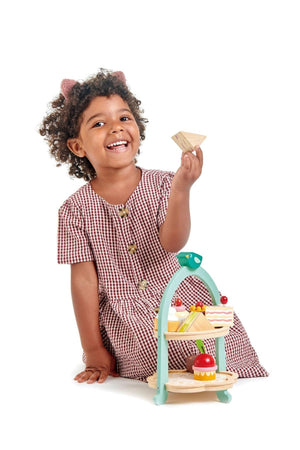 A cheerful child in a gingham dress holds a wooden sandwich while sitting next to the Tender Leaf Wooden Toys Afternoon Tea Stand.