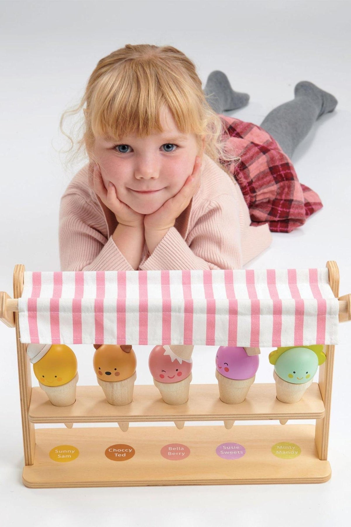 A child lies on the floor, resting her chin on her hands, next to a wooden ice cream shop with.