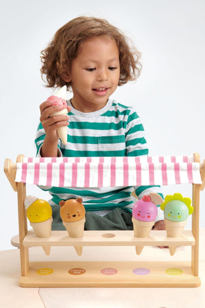A child with curly hair holds a toy ice cream cone, playing at a wooden ice cream shop with colorful.