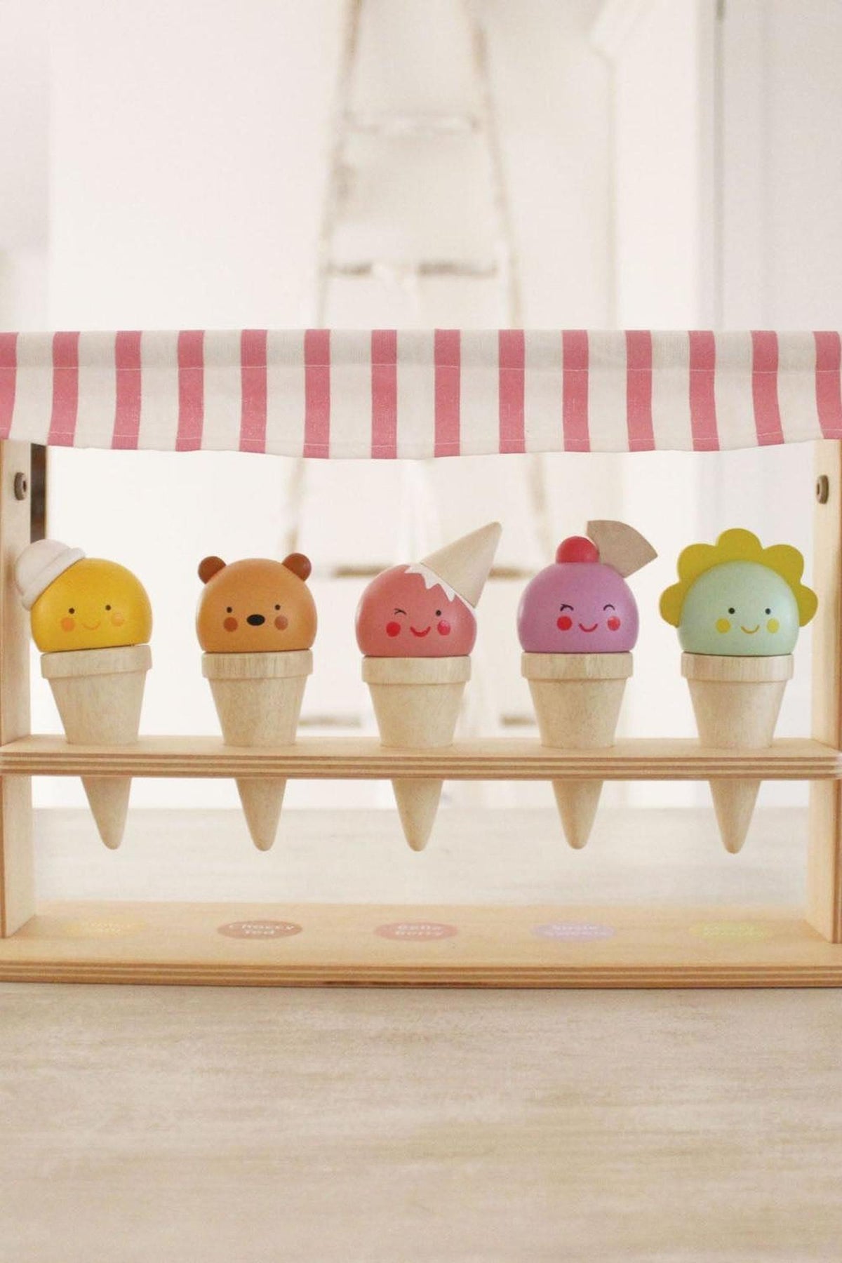Colorful wooden ice cream cones with friendly faces are displayed under a striped pink canopy, showcasing various playful flavors.