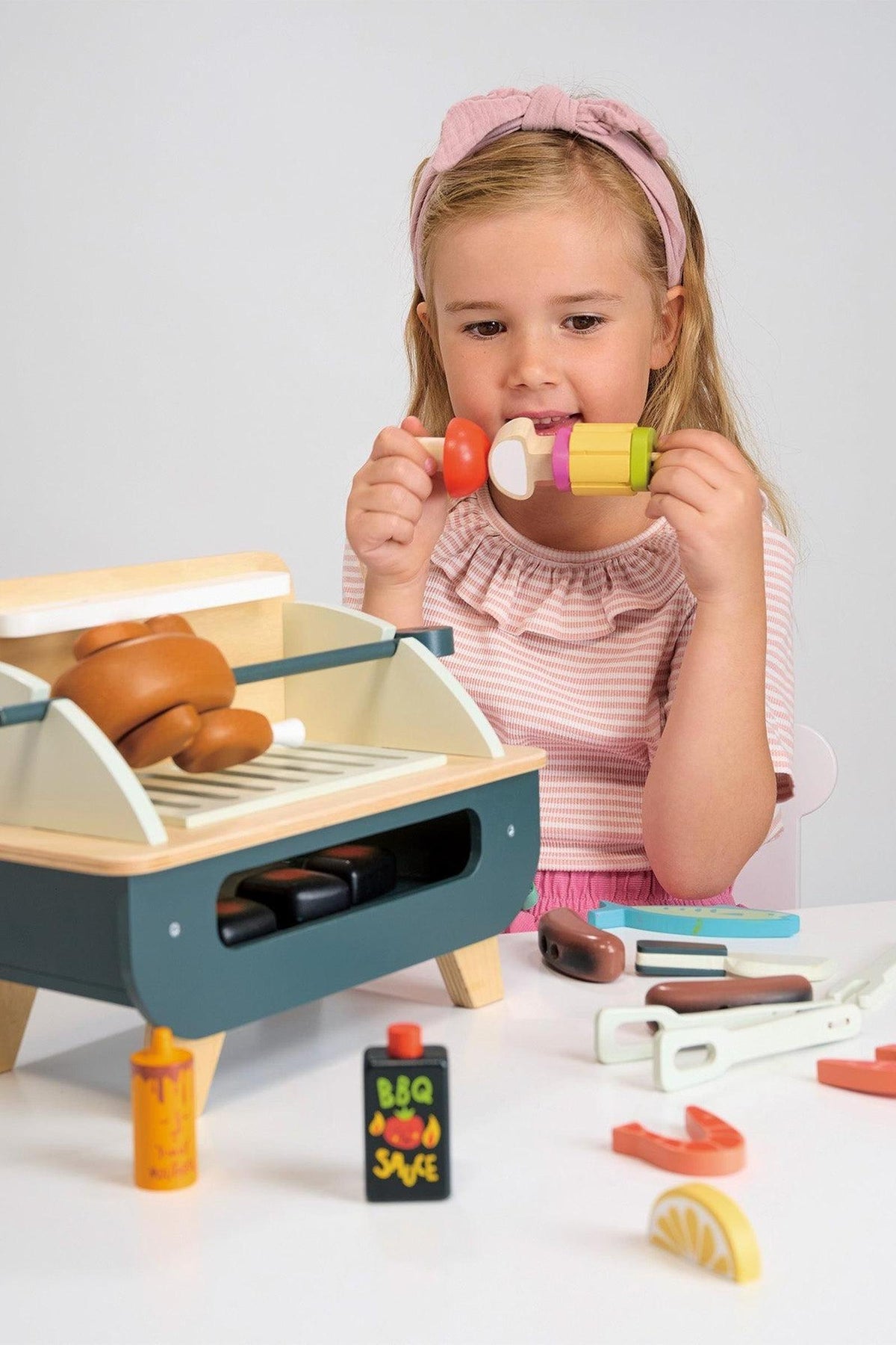 A young girl in a pink top and headband holds colorful wooden food items while interacting with a wooden barbeque playset. The set features a grill, sausages, and condiment bottles.