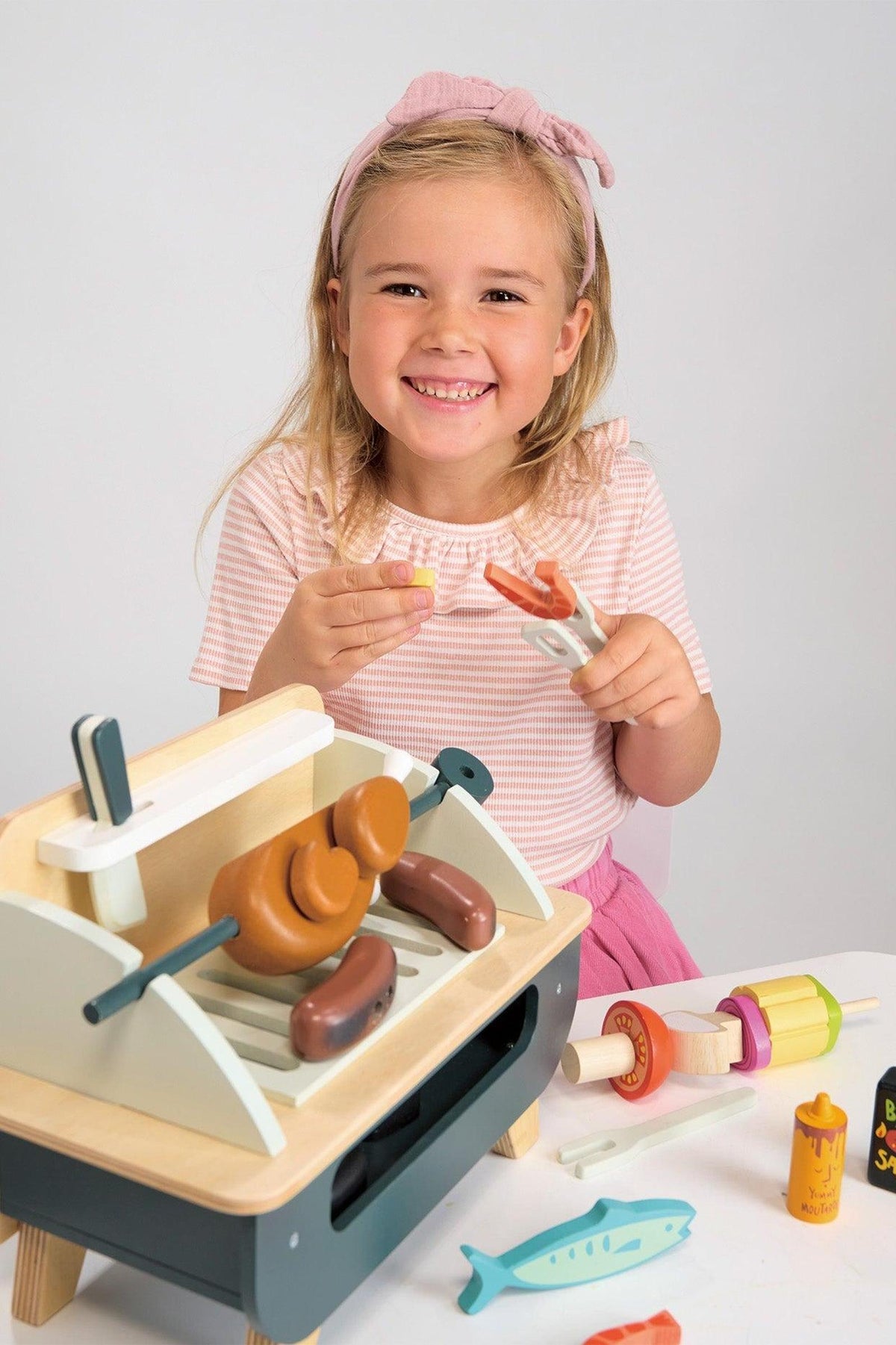 A young girl smiles while holding a wooden sausage and tongs near a colorful wooden barbeque play set with various food accessories, promoting imaginative play and eco-friendly fun.