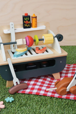 A wooden barbeque play set features a removable grill with colorful, toy food items, including sausages, vegetables, and sauce bottles, set on a checkered picnic blanket. Ideal for imaginative play.