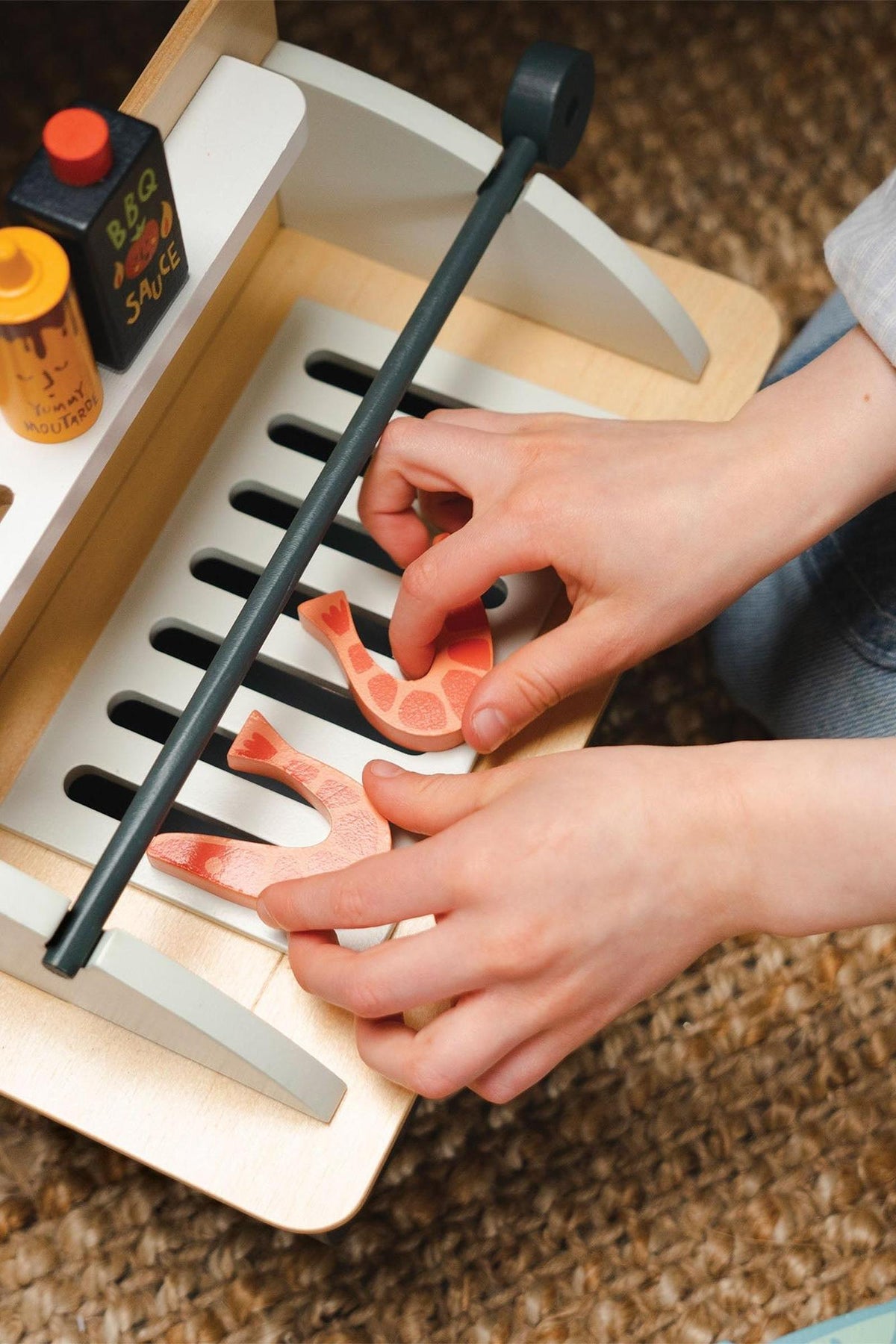 A child’s hands arrange colorful wooden prawns on a toy barbeque grill. The set includes sauce bottles, highlighting its eco-friendly wooden construction and engaging design for imaginative play.
