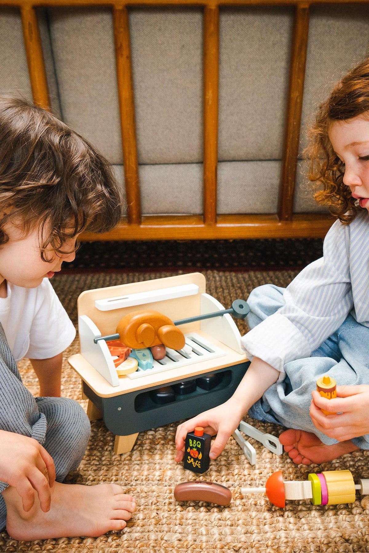 Two children play together with a wooden barbeque toy set, featuring realistic grill accessories, including sausages and condiments. The cozy setting highlights the eco-friendly design of the toy.