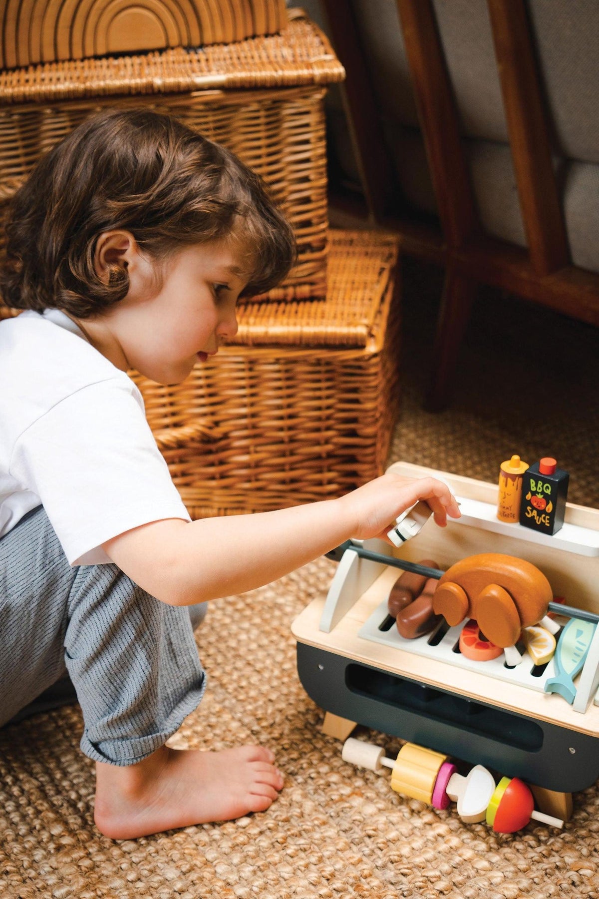 A child with curly hair plays with a wooden barbeque set, featuring various colorful accessories like sausages, vegetables, and sauce pots.