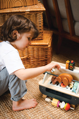 A child with curly hair plays with a wooden barbeque set, featuring various colorful accessories like sausages, vegetables, and sauce pots.