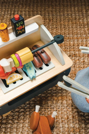 A colorful wooden barbeque play set features various accessories, including sausages, fish, and vegetable pieces, alongside a bottle of BBQ sauce, promoting imaginative play for children.