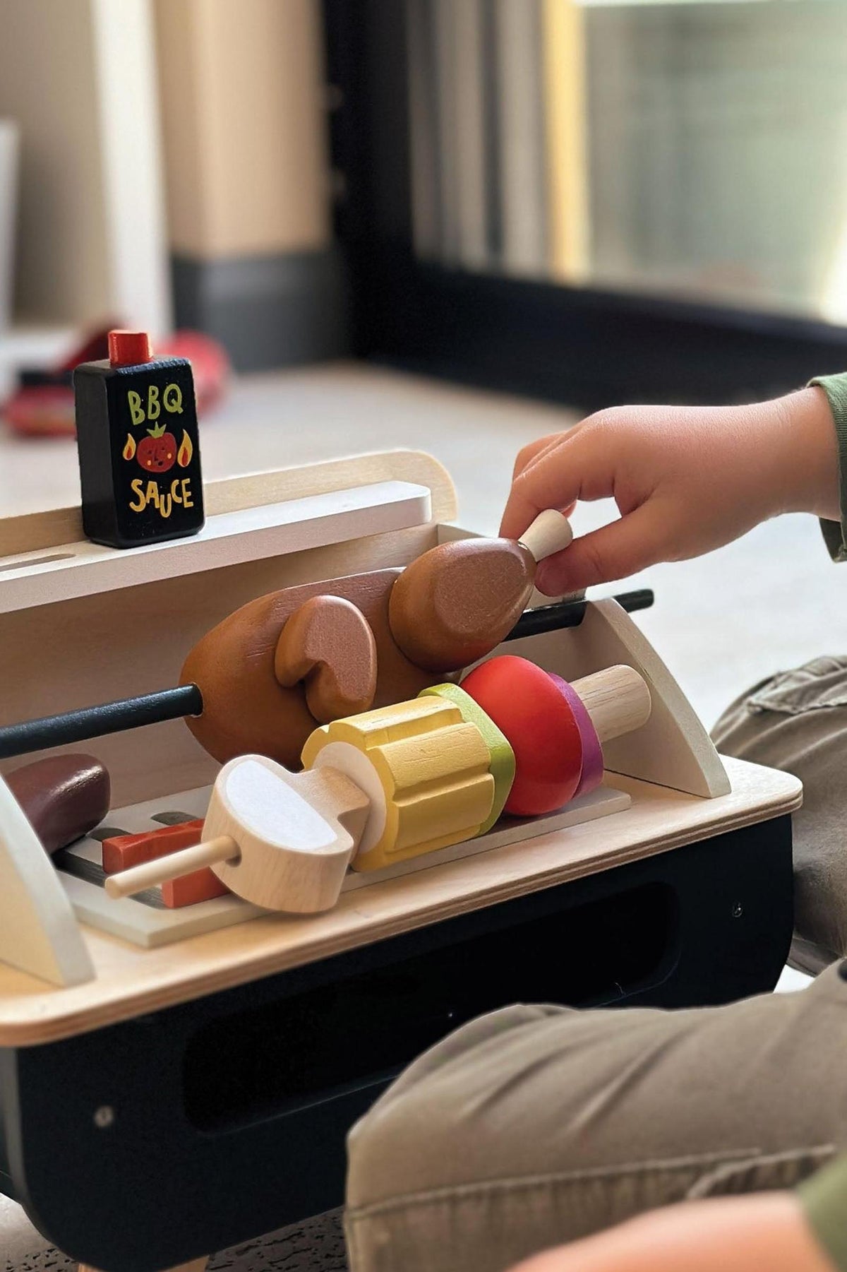 A child’s hand reaches for colorful wooden play food, including chicken and vegetable pieces, on a self-standing wooden barbeque toy.