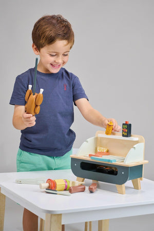 A young boy smiles while holding wooden tongs and play food near a self-standing wooden barbeque toy. The set features colorful accessories, promoting imaginative play and sustainable materials.