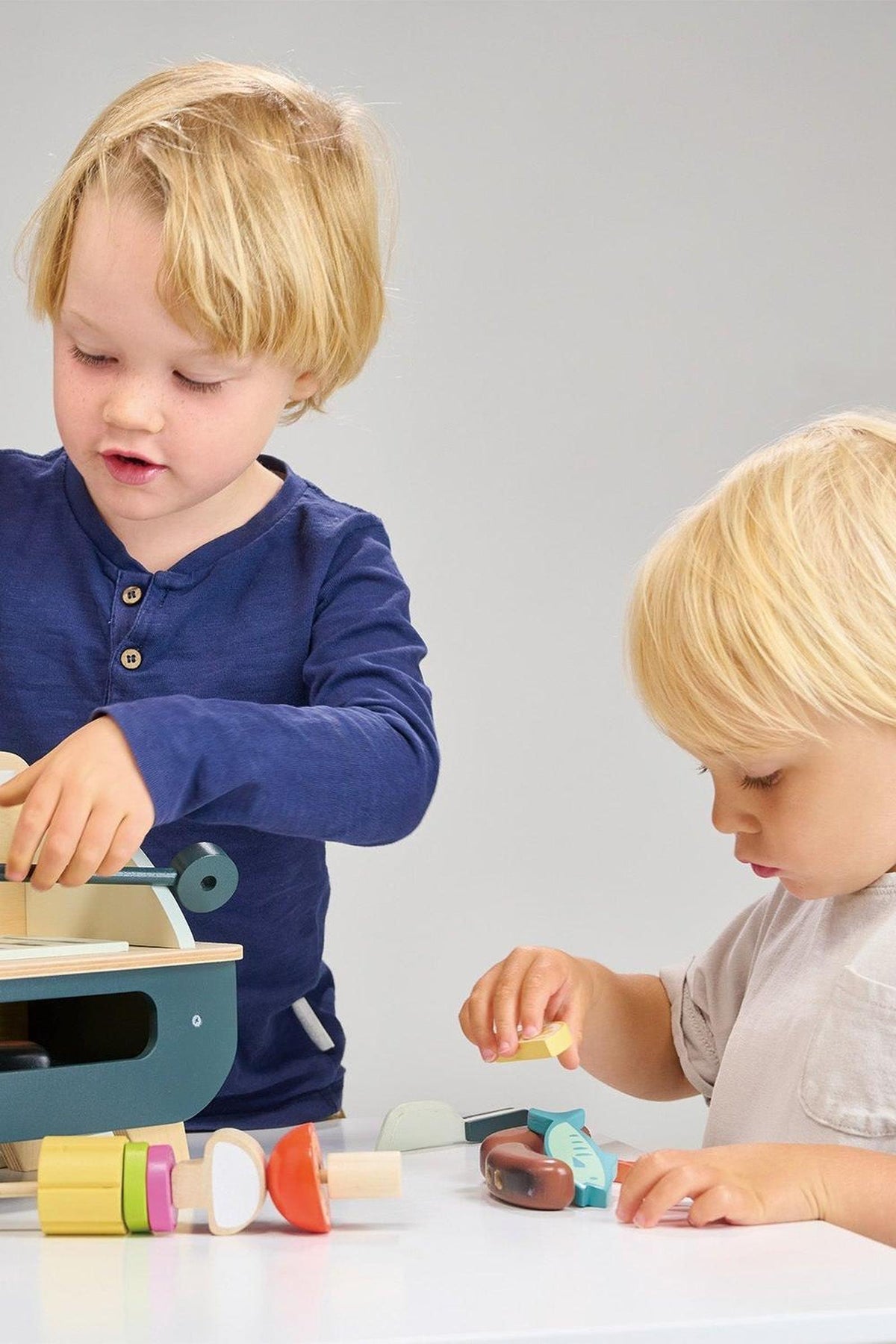 Two children play with a wooden barbeque set, featuring colorful, sustainable accessories. One child interacts with the grill while the other examines various food pieces, promoting imaginative play.