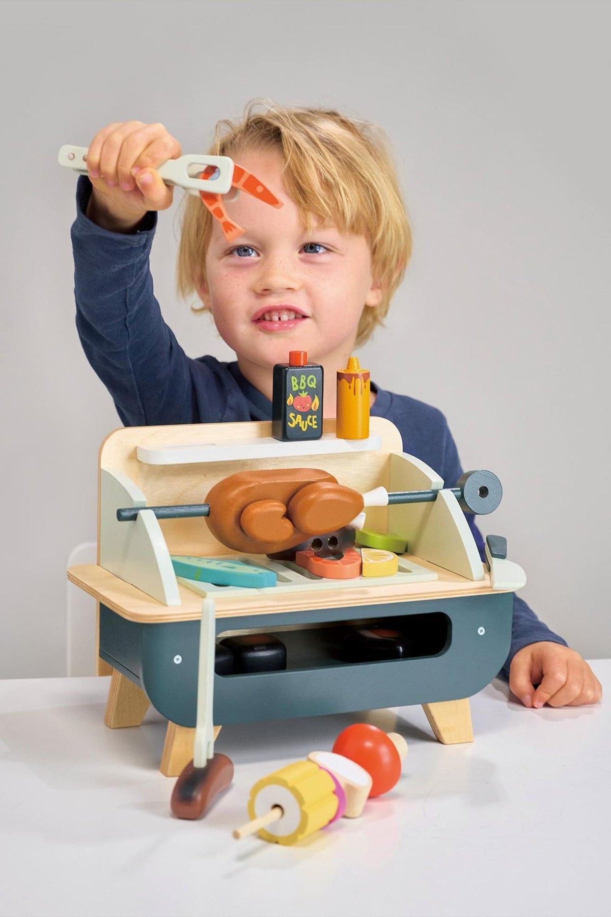 A young child joyfully plays with a wooden barbeque toy set, featuring a rotisserie chicken, sausages, and colorful vegetable pieces, showcasing eco-friendly craftsmanship in a bright, inviting setting.