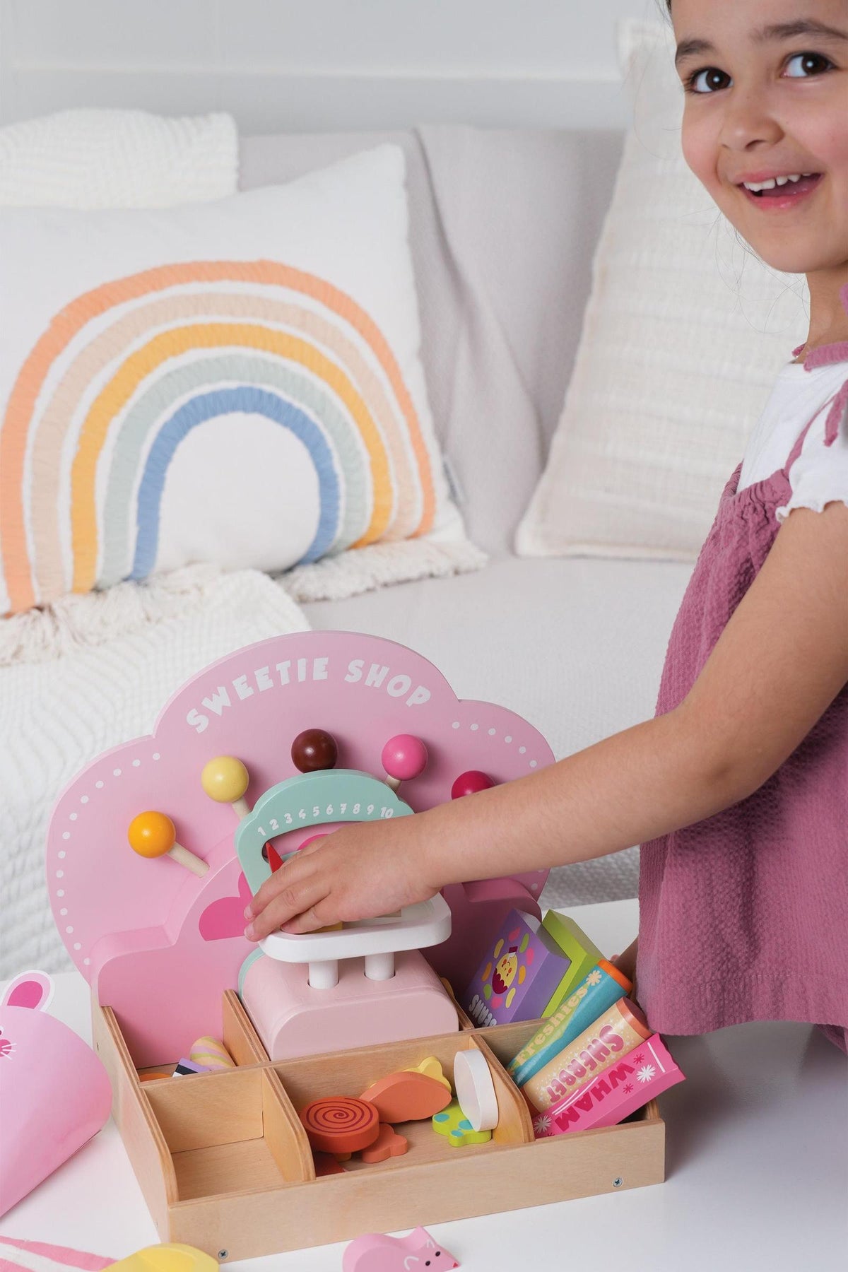 A young child joyfully plays with the Tender Leaf Wooden Toys Sweetie PicnMix Shop, featuring colorful candies and a decorative scale. The shops charming design encourages imaginative play.