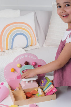 A young child joyfully plays with the Tender Leaf Wooden Toys Sweetie PicnMix Shop, featuring colorful candies and a decorative scale. The shops charming design encourages imaginative play.