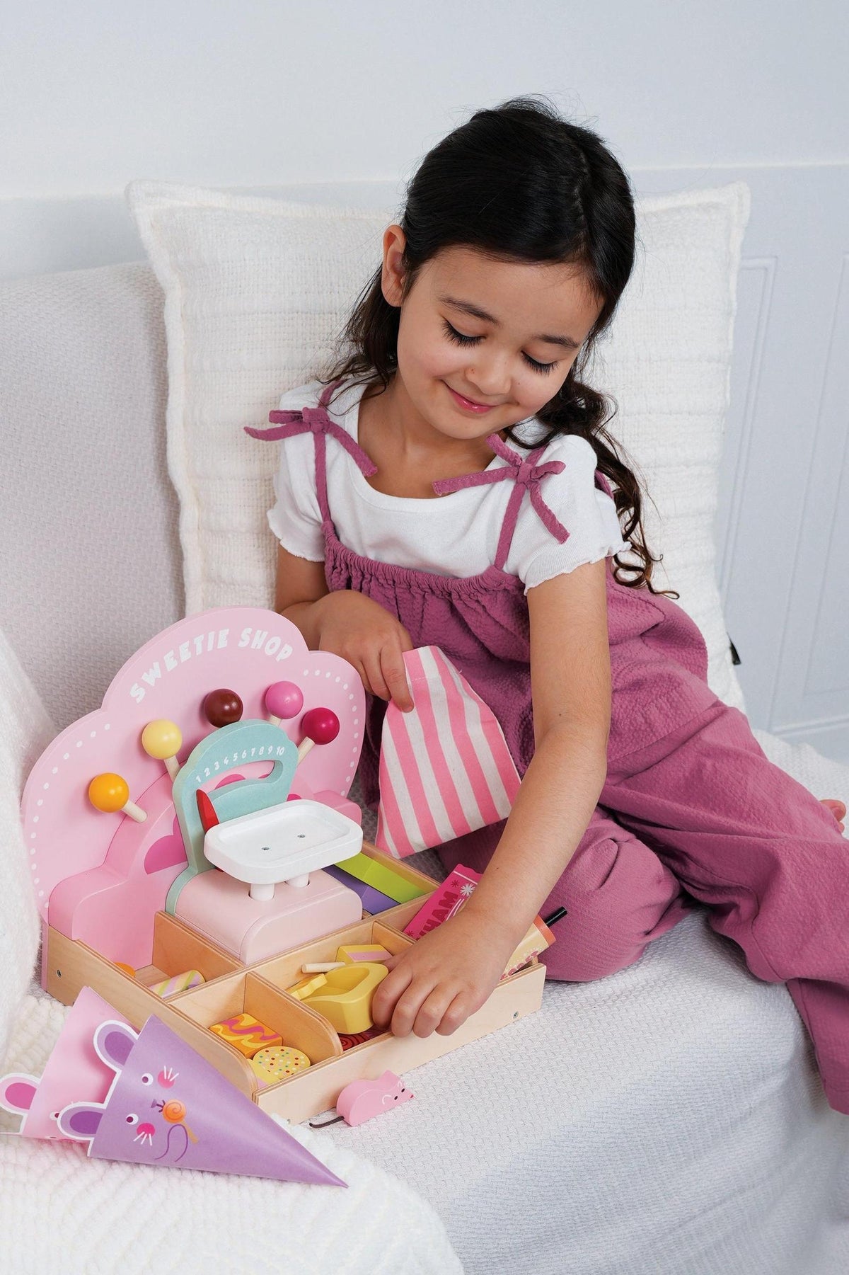 A young girl sits on a white sofa, joyfully playing with the Tender Leaf Wooden Toys Sweetie PicnMix Shop, featuring colorful candies and a decorative display.