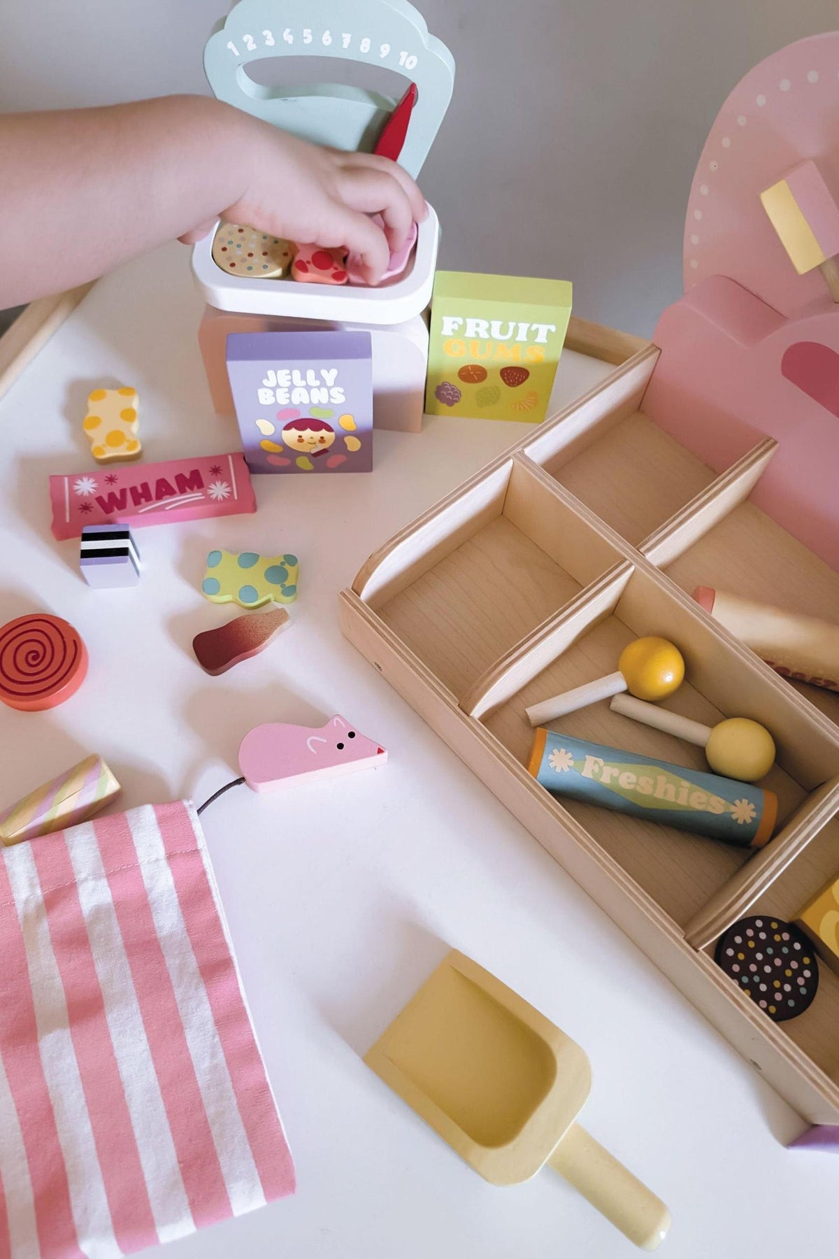 A child’s hand reaches into a colorful wooden sweet shop playset, featuring various candies, a scoop, and labeled compartments. The scene is vibrant and inviting, encouraging imaginative play.