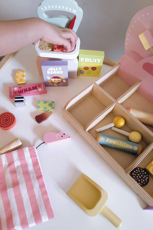 A child’s hand reaches into a colorful wooden sweet shop playset, featuring various candies, a scoop, and labeled compartments. The scene is vibrant and inviting, encouraging imaginative play.