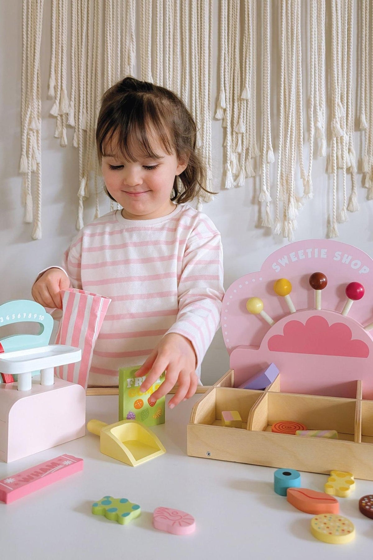 A young child in a striped pink and white shirt plays with a colorful wooden Sweetie PicnMix Shop toy, featuring various candy-themed pieces and a decorative shelf in a bright.