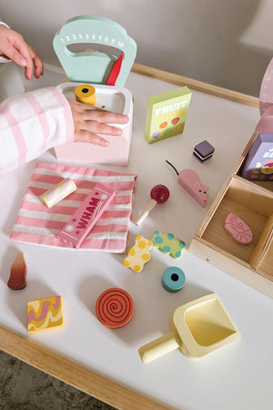 A childs hand reaches for a colorful wooden scoop beside a scale, surrounded by various playful wooden treats and a pink striped cloth, enhancing imaginative play with Tender Leaf Toys.