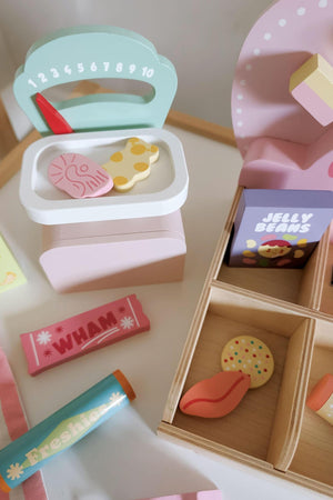 A colorful wooden toy set featuring a pastel pink cash register, various candy-themed pieces, and compartments filled with sweets like jelly beans and a chocolate bar, designed for imaginative play.