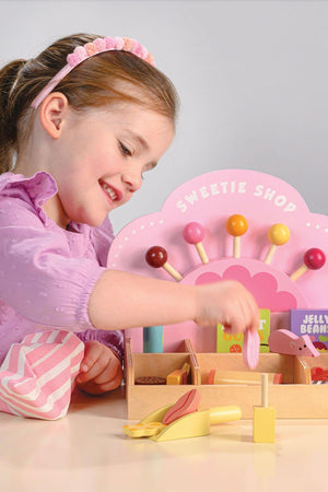 A young girl in a purple shirt plays with the Tender Leaf Wooden Toys Sweetie PicnMix Shop, using a scoop to select colorful wooden candies from the shops compartments.