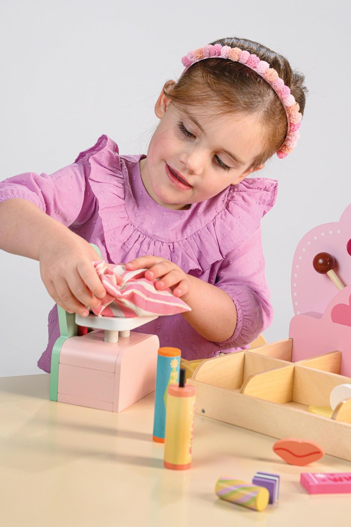 A young girl in a pink blouse uses a toy scoop to select colorful candies from a wooden sweet shop playset.