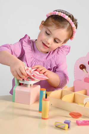 A young girl in a pink blouse uses a toy scoop to select colorful candies from a wooden sweet shop playset.